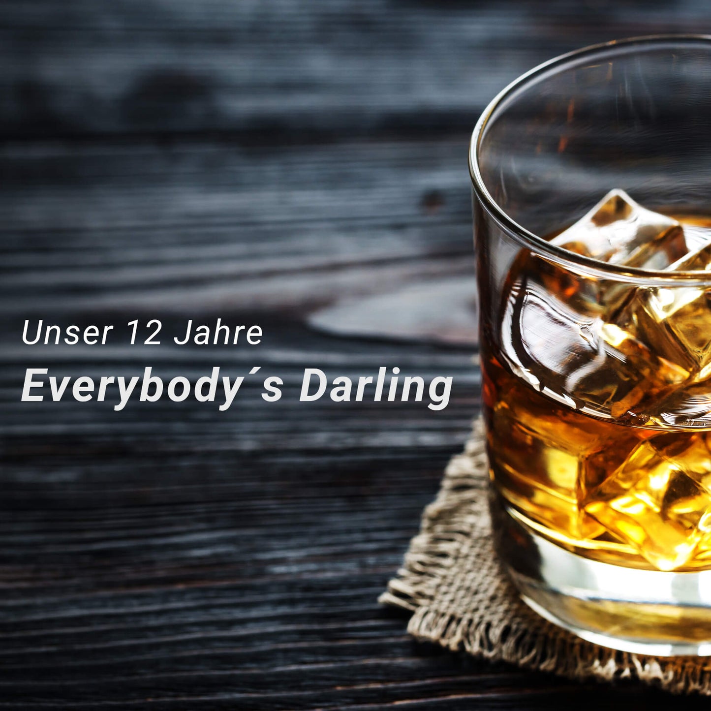 Tumbler Glas mit Rum auf einem schwarzen Holztisch stehend und der Schrift "Unser 12 Jahre, Everybody´s Darling"